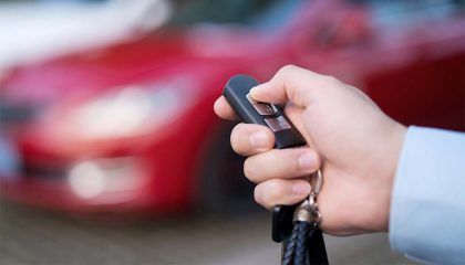 A person holding a black car key fob in front of a blurred red vehicle.